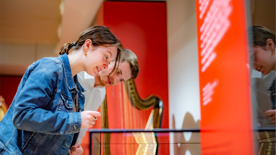 Two visitors to the Royal College of Music Museum look downwards into a glass showcase containing items on display. A harp is placed in the background.